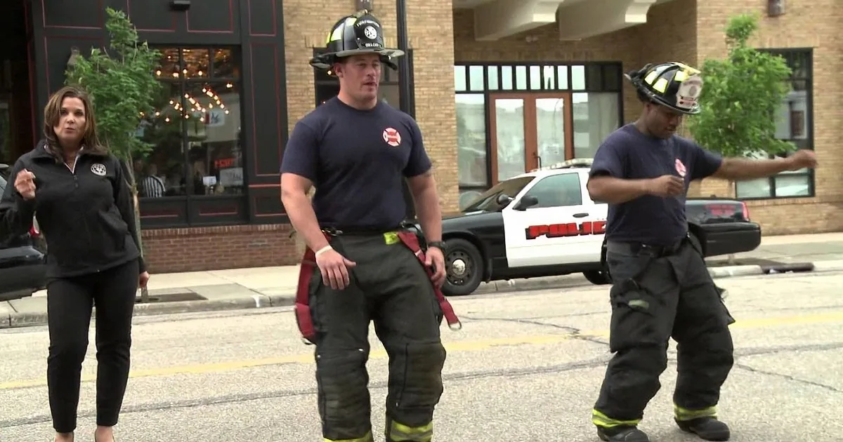 Two firefighters in gear stand on a street, while a woman in black observes nearby.