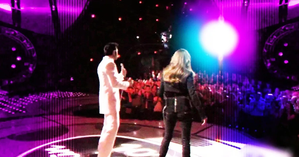 A man in white and a woman in black stand on a stage with vibrant purple and blue lighting, facing each other.