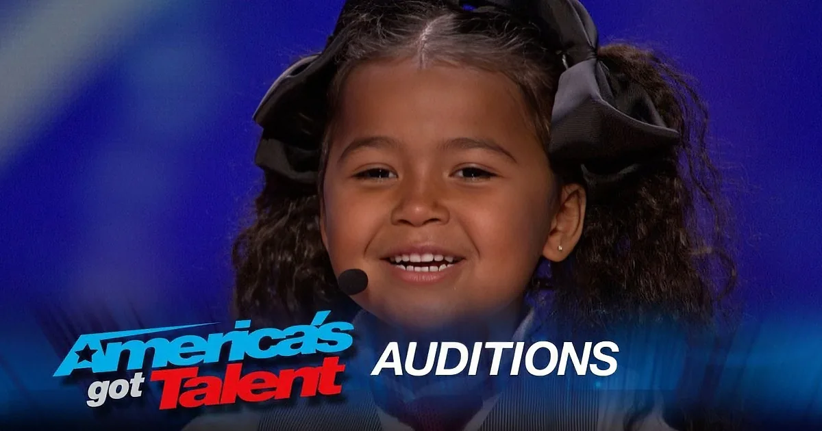 A young girl with curly hair smiles on stage at America's Got Talent auditions.