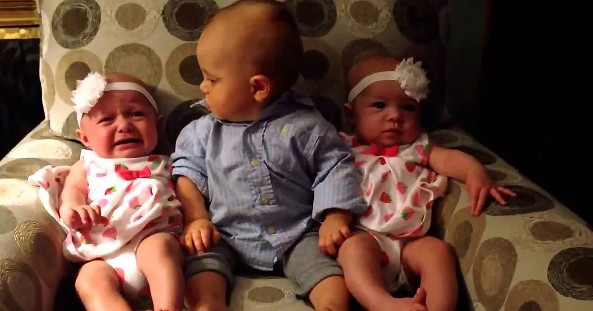 Three babies, two in matching outfits, sit on a patterned couch, one crying while the others look on.