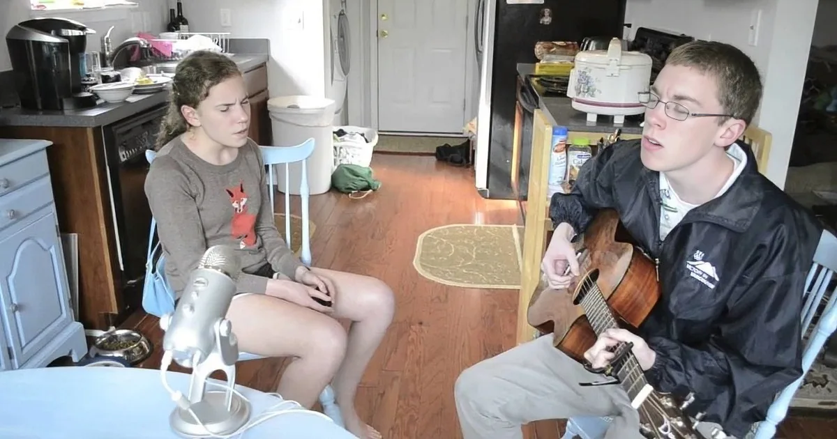 A man plays guitar while a woman sits nearby, both in a kitchen setting with various household items visible.