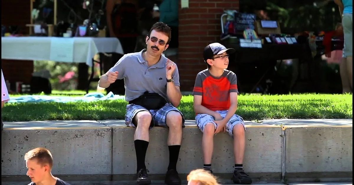 A man and a boy sit on concrete steps, the man gesturing with his hands, both wearing casual summer attire.