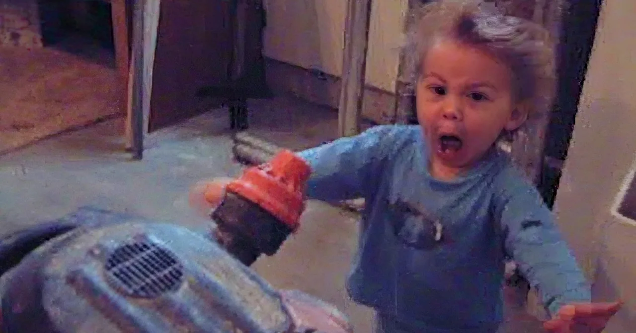 A toddler in a blue shirt is sitting on a toy motorcycle, appearing excited.