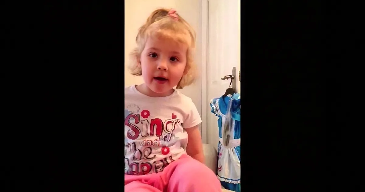 A young child with blonde hair in a ponytail, wearing a white shirt with red text and pink pants, sits in a room with...