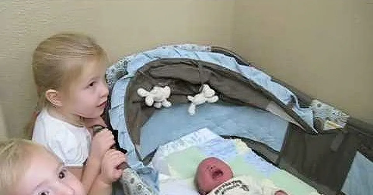 A young child gazes at a baby in a crib, surrounded by stuffed animals and a blanket.