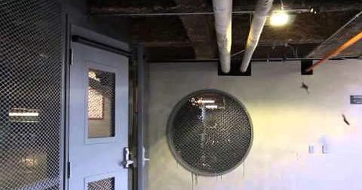 A circular vent in a wall with a door and mesh screen nearby, pipes above, and a light fixture.