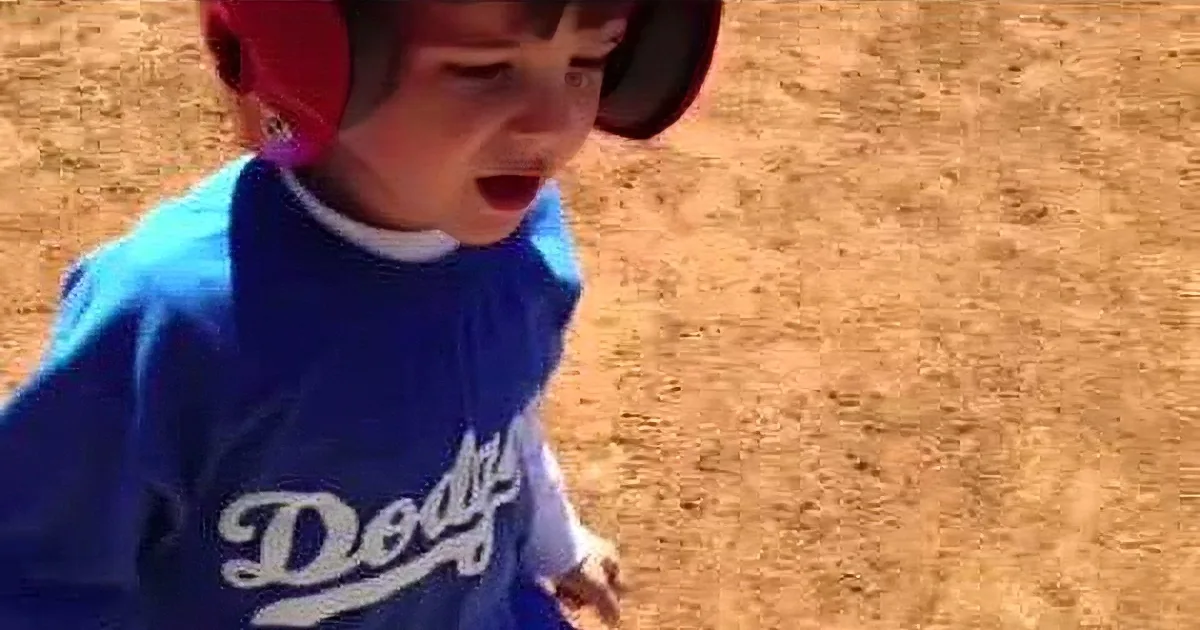 A young boy in a blue Dodgers jersey and red ear protectors stands on a dirt field, looking down.