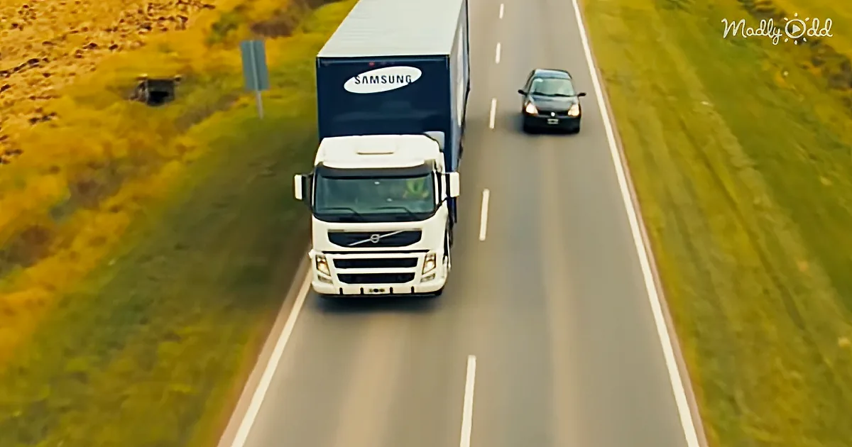 A white Volvo truck with a Samsung trailer drives on a highway with a black car behind it.