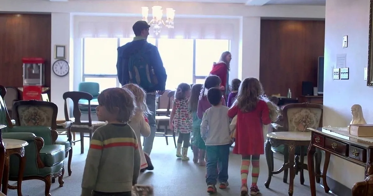 A group of children in colorful outfits walks through a warmly lit room with vintage furniture and a chandelier.