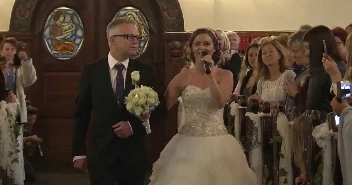 A bride in a white gown walks down the aisle with a man in a suit, holding a microphone, as guests watch.