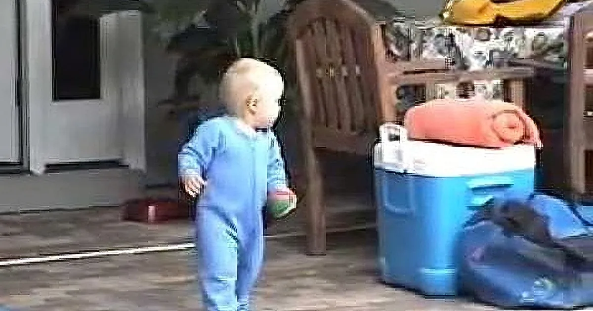 A baby in blue pajamas walks on a patio near a cooler and outdoor furniture.