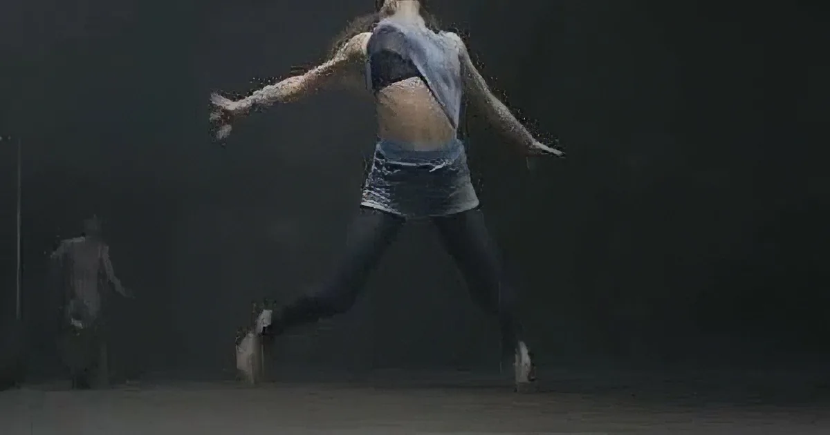 A dancer in mid-air, wearing a blue top and black pants, with arms outstretched and a dark background.