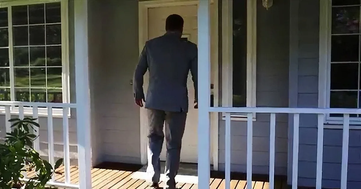 A man in a suit walks up the steps of a house with white railings and a wooden deck.