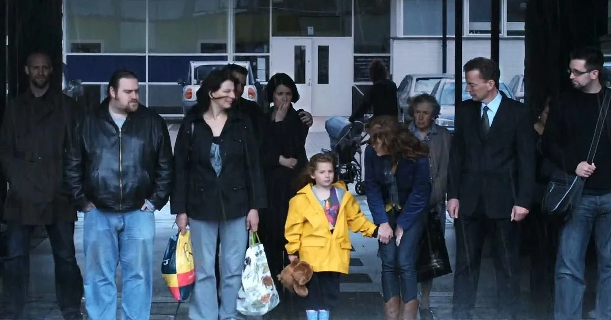 A group of people, including a child in a yellow jacket, walks outside a building on a rainy day.