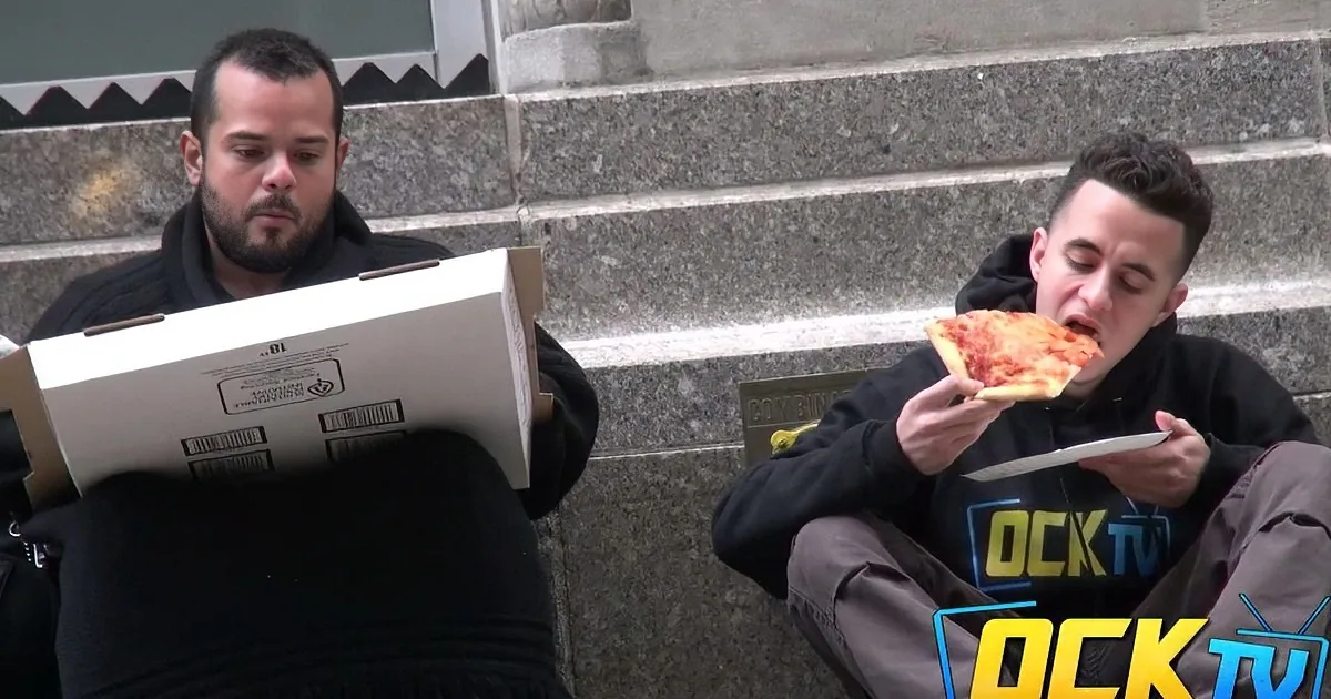 Two men sit on steps, one eating pizza, the other holding a pizza box.