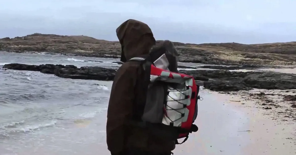 A person in a hooded jacket and backpack stands on a rocky beach, facing the ocean under a cloudy sky.