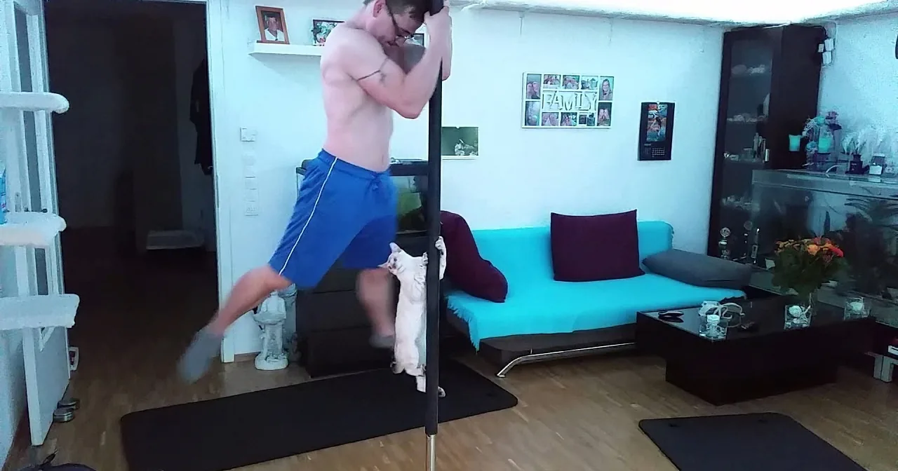A shirtless man in blue shorts is doing a pull-up exercise in a living room with a couch and a coffee table.