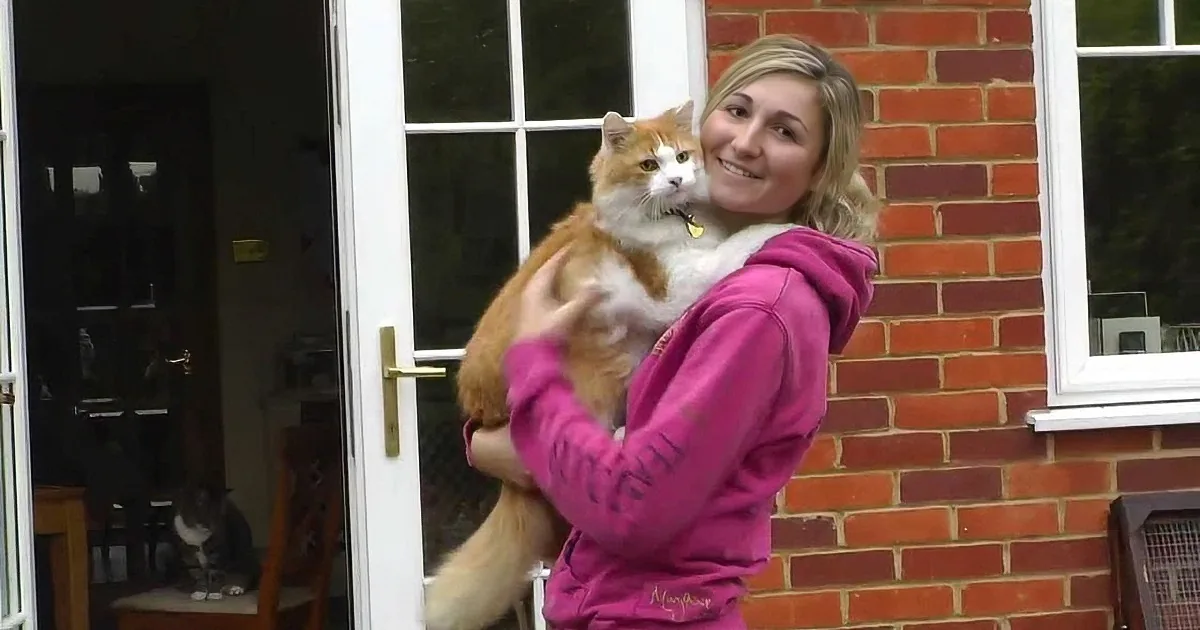 A woman in a pink hoodie holds a large orange and white cat outside a brick house.