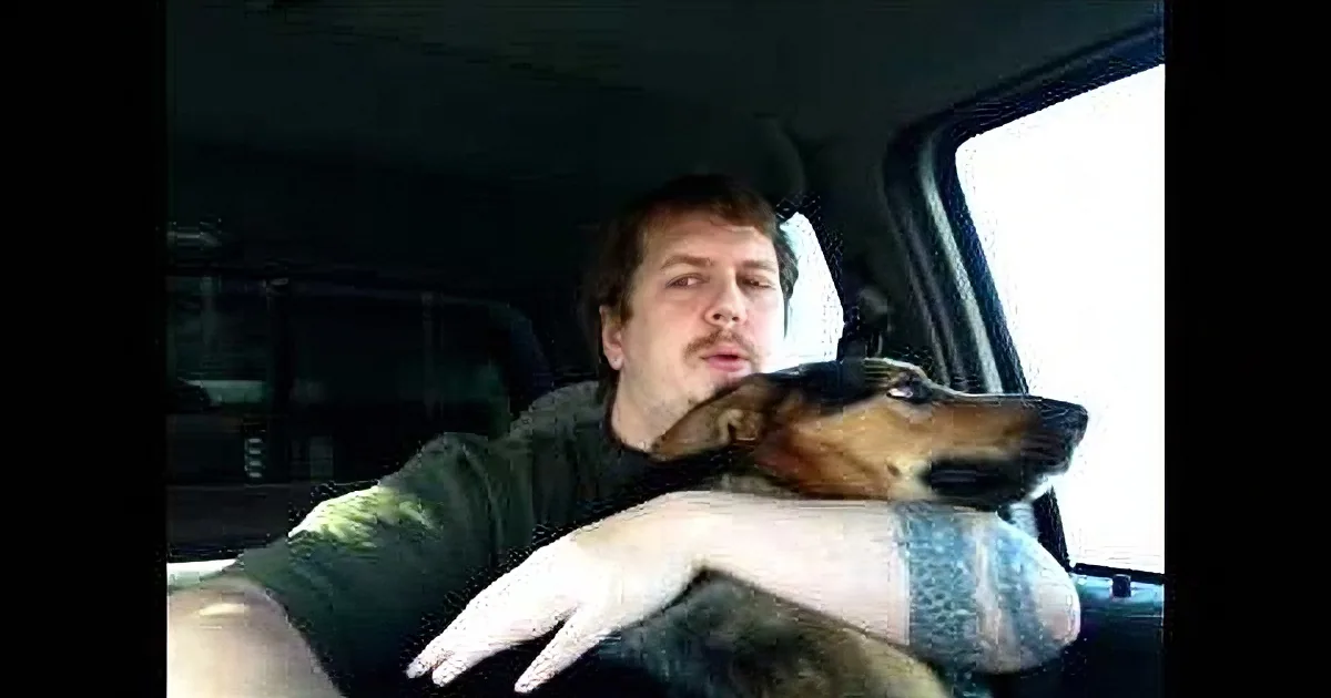 A man sits in a car with a dog resting its head on his shoulder, both looking out the window.
