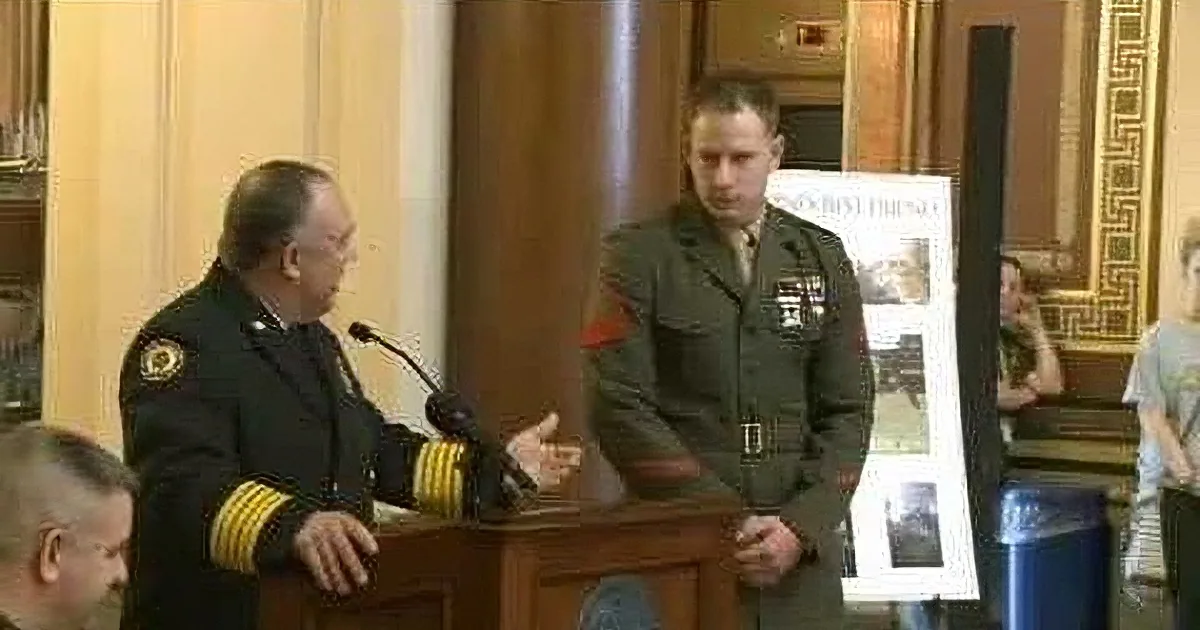 A man in a military uniform stands at a podium, addressing an audience, with another in uniform beside him.