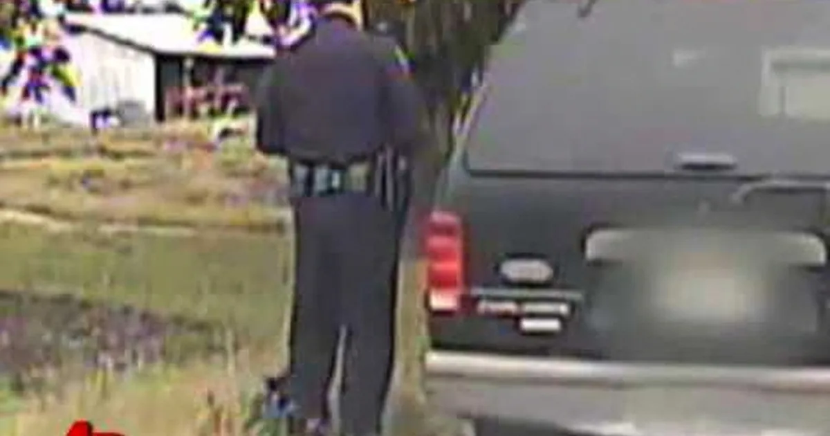 A person in dark clothing walks away from a vehicle in a grassy area near a house.