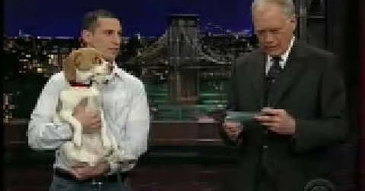 A man holds a dog while another man in a suit looks on, both on a talk show set with a cityscape backdrop.