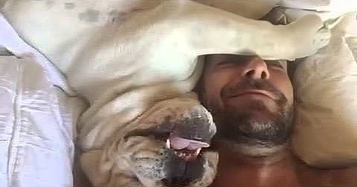 A man is lying in bed with a large, light-colored dog resting its head on his chest.