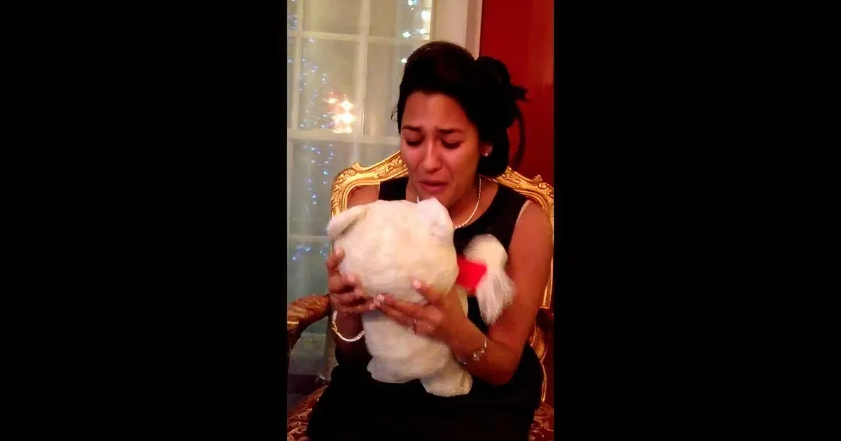 A woman in a black dress holds a white teddy bear, appearing emotional, with a decorated window in the background.