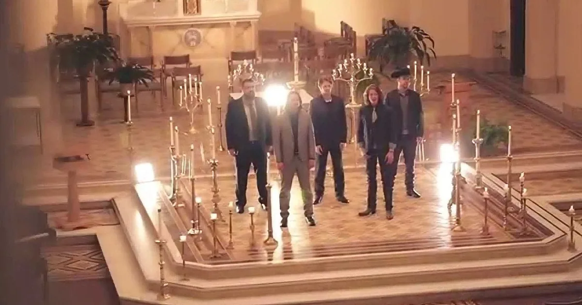 Six people in formal attire walk down a candle-lit aisle in a grand, ornate church.