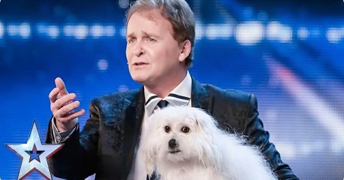 A man in a suit holds a white dog, gesturing with his hand, on a stage with a star logo.