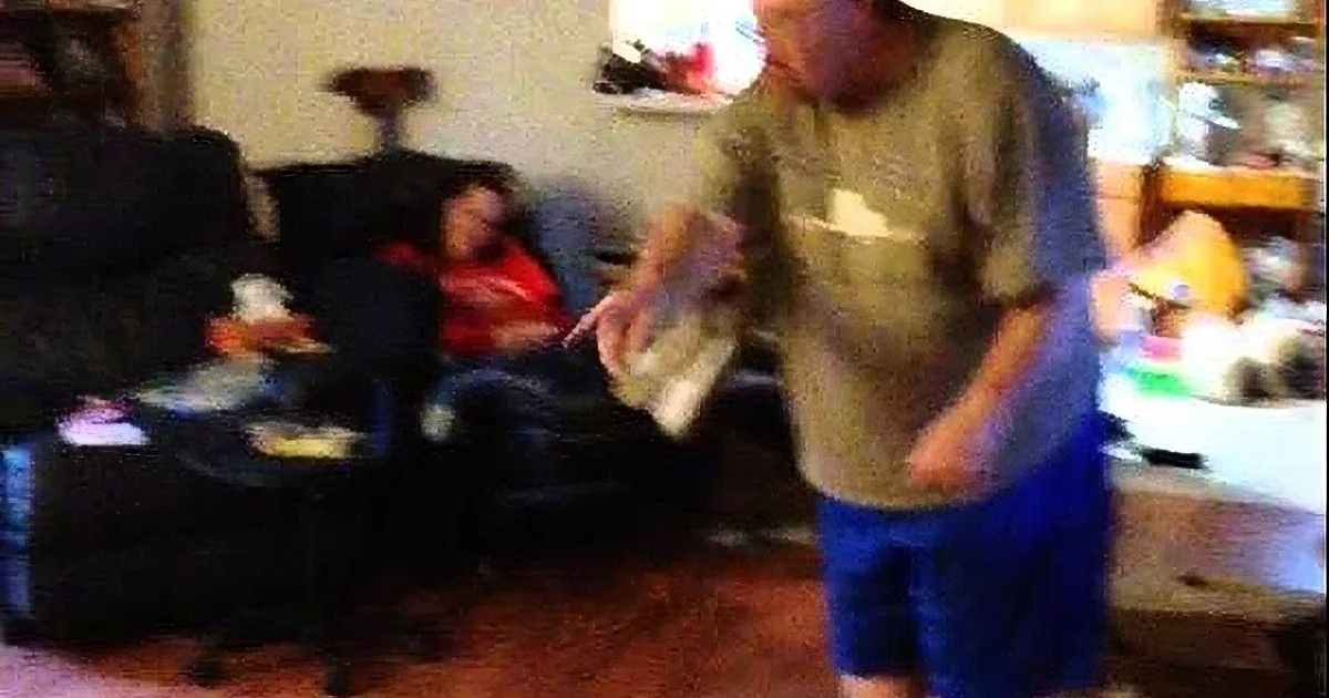 A man in a gray shirt and blue shorts stands in a living room, holding a white object, while a woman sits on a black...