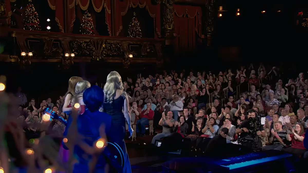 A performer in a blue costume stands on stage, facing a large audience applauding in a festive theater setting.