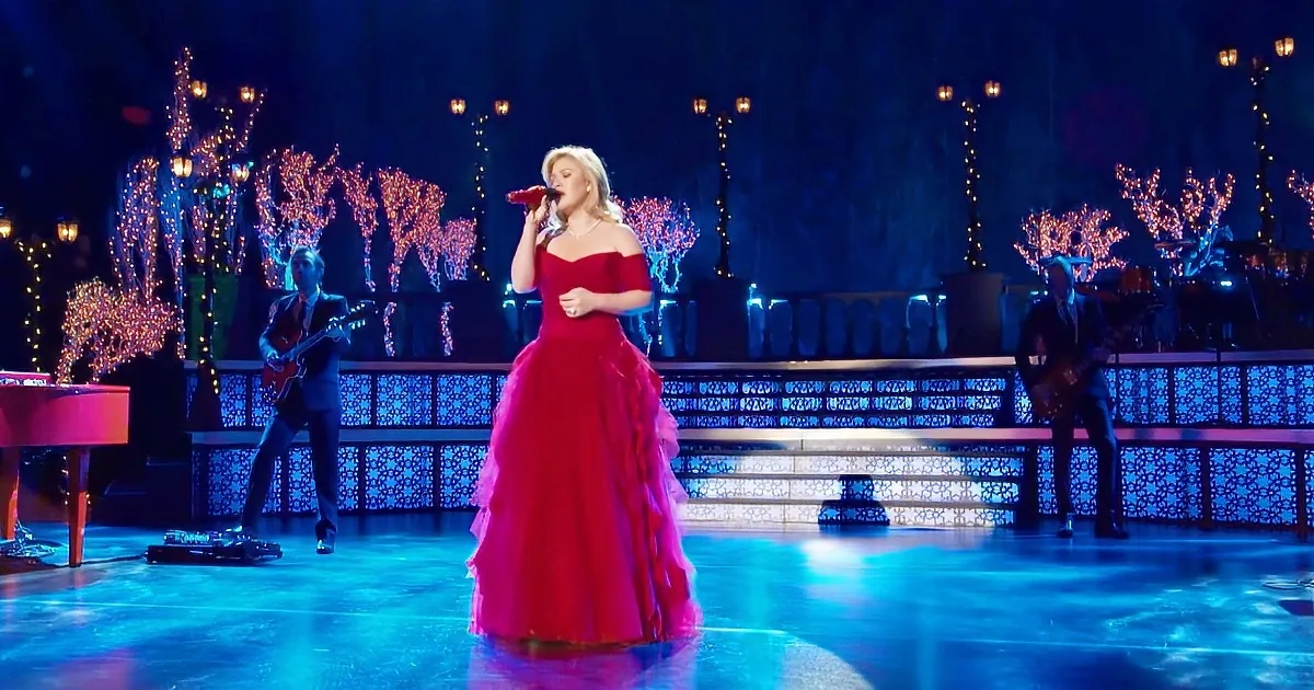 A woman in a red dress sings on stage with musicians and illuminated trees in the background.
