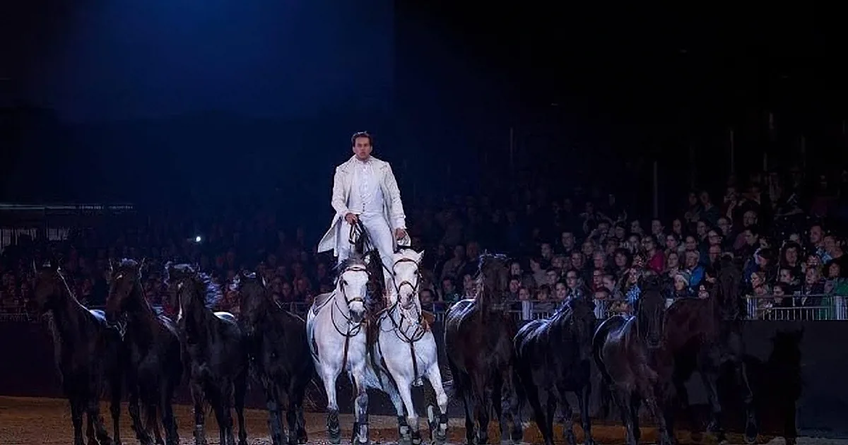 A man in white attire rides a white horse amidst a group of black horses in a dimly lit arena.