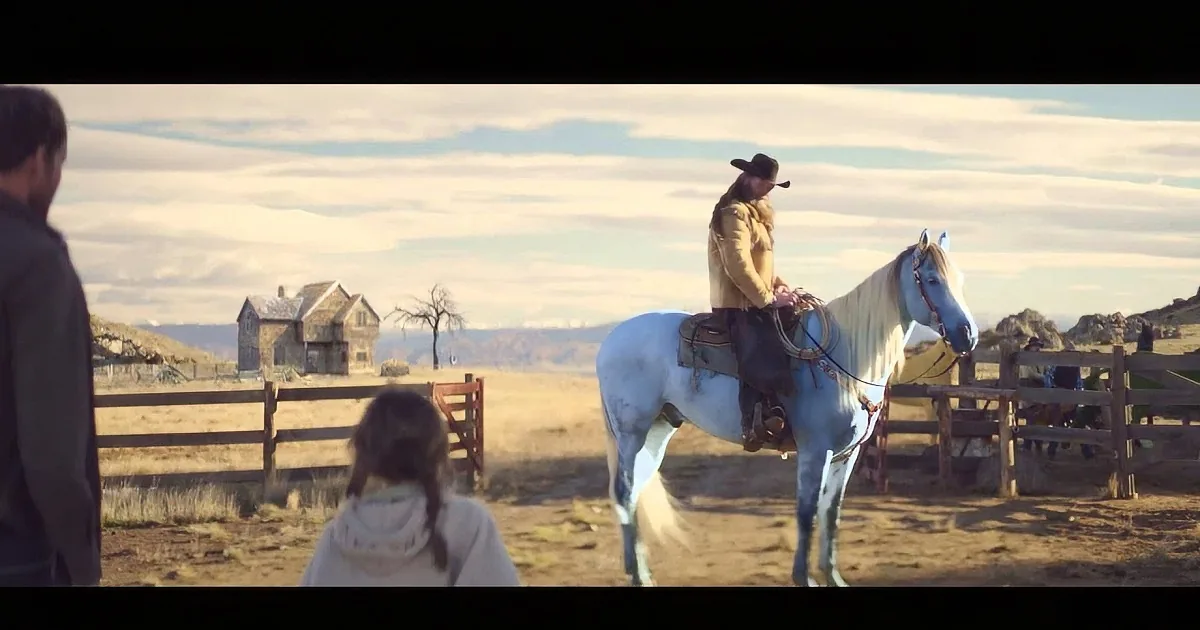 A man in a cowboy hat rides a white horse near a wooden fence and a house in a rural setting.