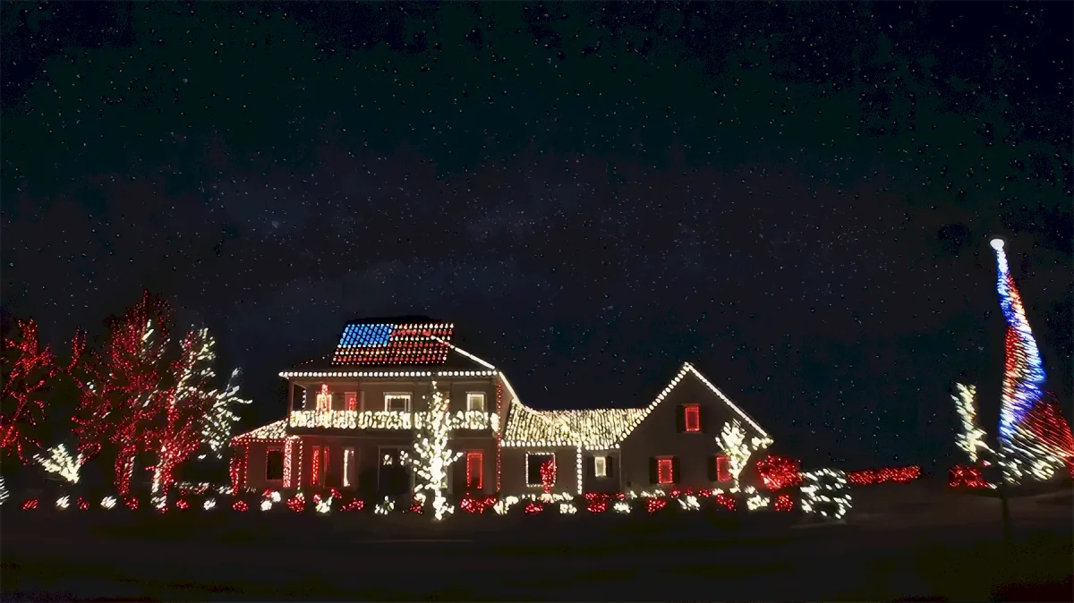 A house adorned with festive lights and an American flag on its roof stands under a starry night sky.