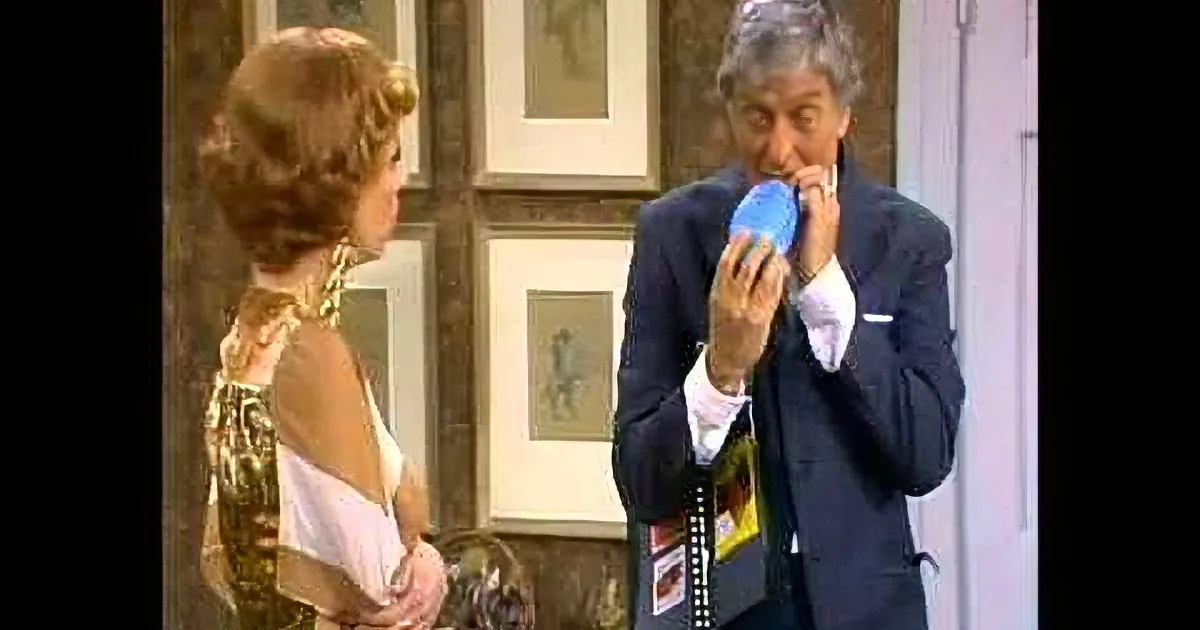 A man in a suit holds a blue object near his mouth, while a woman in a gold dress stands beside him.