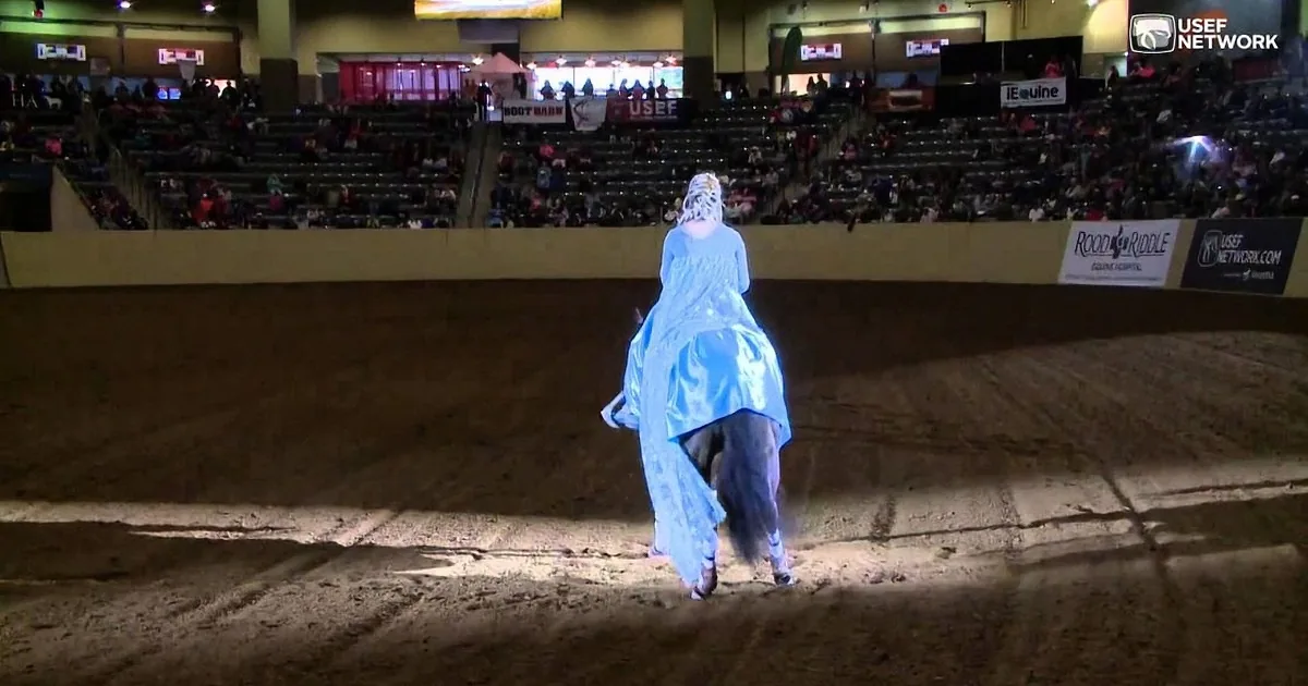 A person in a blue dress rides a horse in an indoor arena, with spectators in the background.