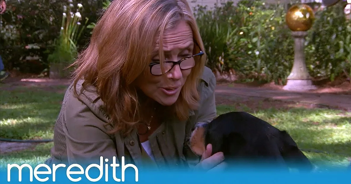 A woman with glasses holds a black dog in a garden setting, with "Meredith" text at the bottom.