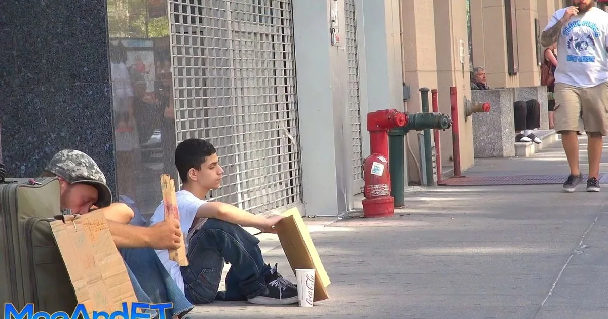 A man sits on a sidewalk holding a cardboard sign, with a Coca-Cola cup nearby.