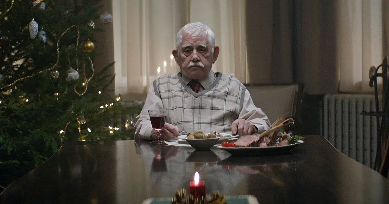 An elderly man sits at a table with a Christmas tree in the background, surrounded by holiday decorations and food.
