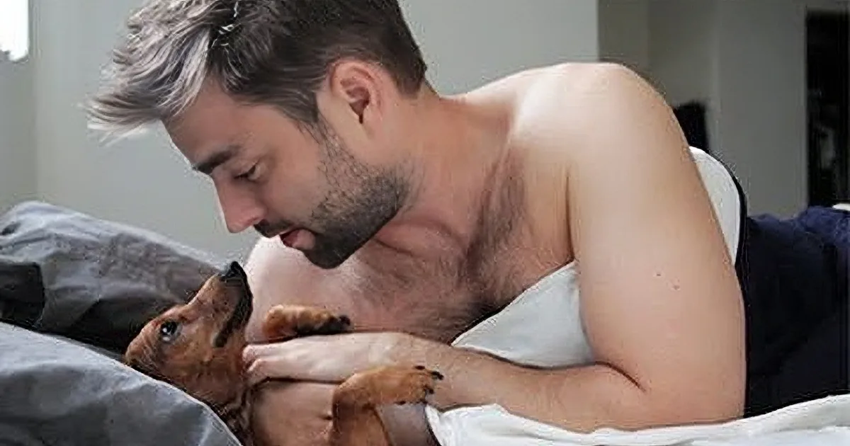 A shirtless man is lying on a bed, holding and cuddling a small brown dog.