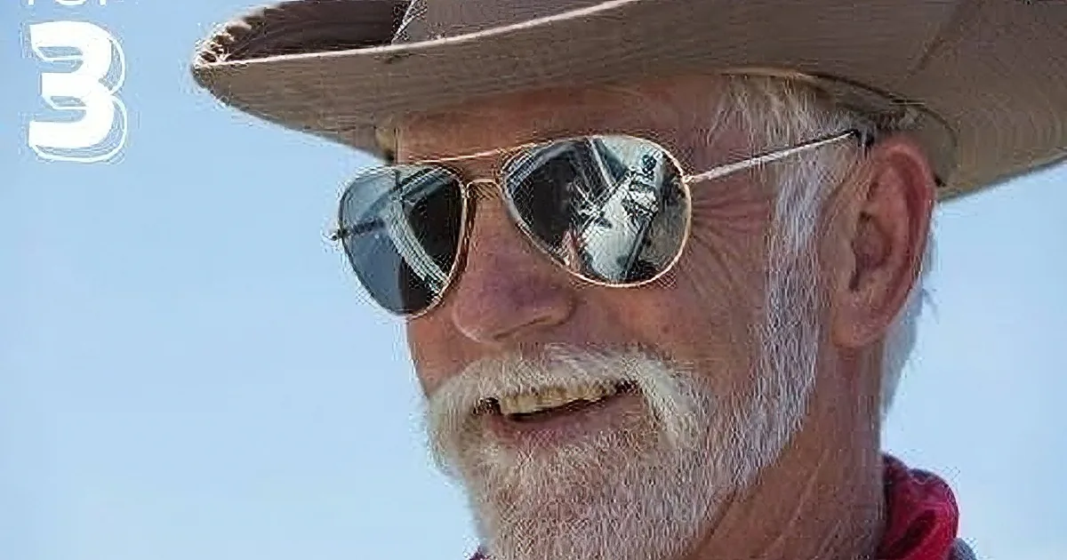 A man with a white beard and sunglasses, wearing a cowboy hat, is smiling against a blue sky.