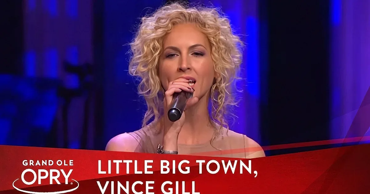 A woman with curly blonde hair sings into a microphone at the Grand Ole Opry.