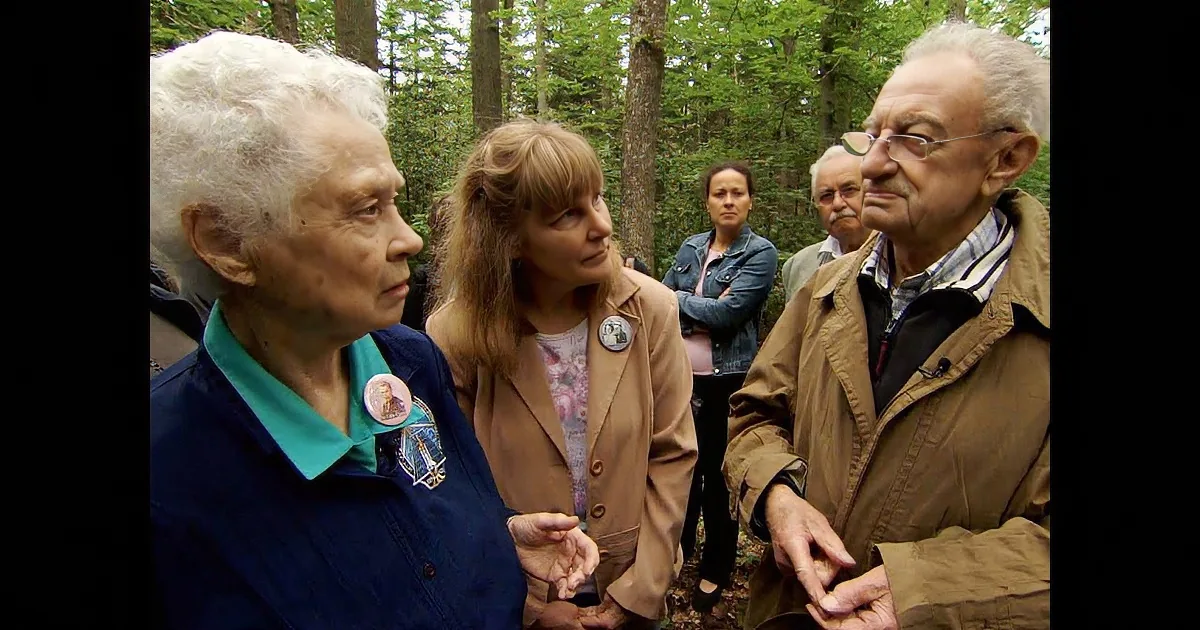 An elderly woman in a blue jacket with a badge talks to a man in a brown jacket in a forest setting.