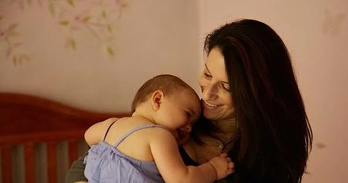 A woman in a blue dress holds a baby close, both smiling warmly in a cozy room.