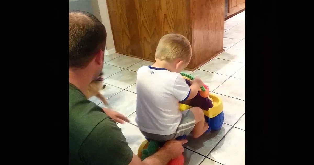 A child sits on a colorful toy car, supported by an adult, in a kitchen setting.
