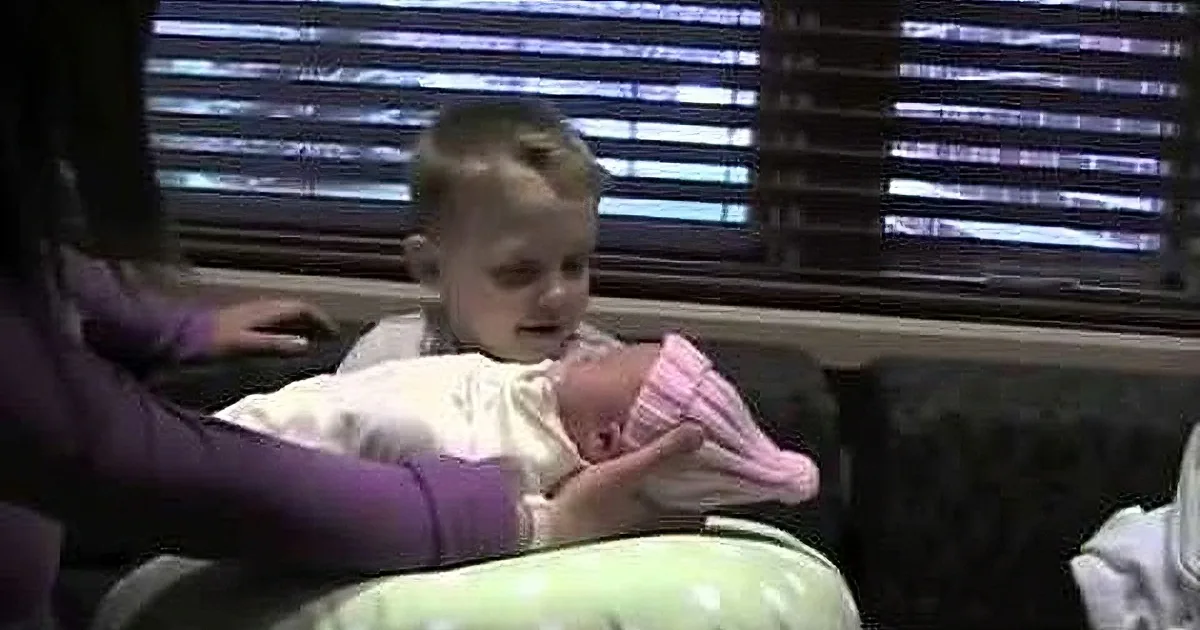 A baby in a pink blanket is being held by a person in a purple shirt, with a window and blinds in the background.