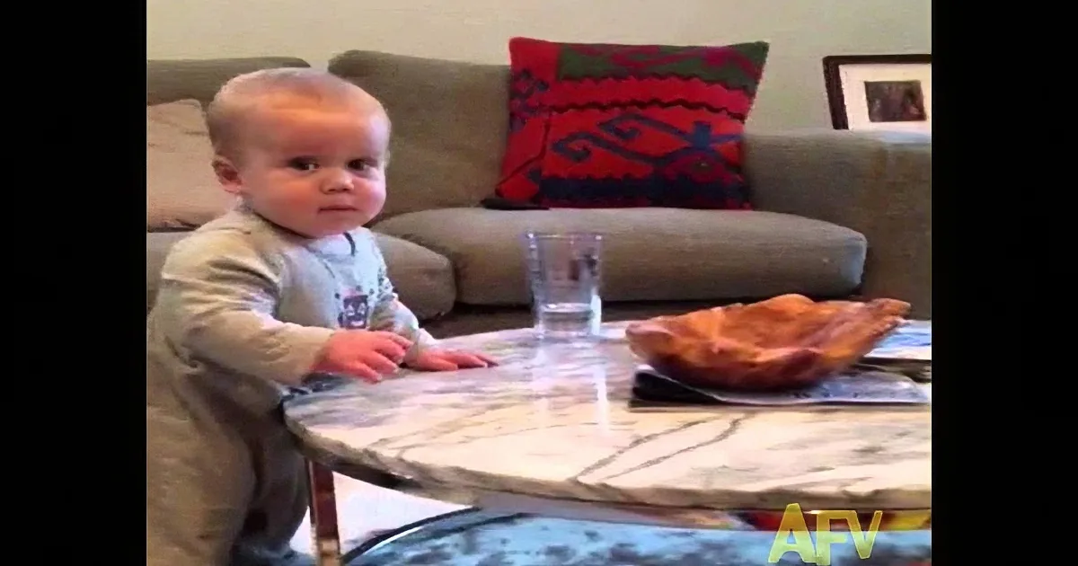 A baby stands at a table with a pizza and a glass of water, looking at something off-camera.