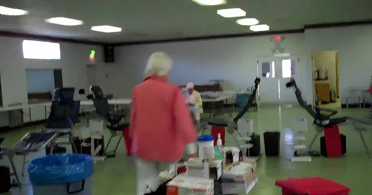 A woman in a pink jacket walks through a room with tables, chairs, and boxes, possibly a community center or event space.